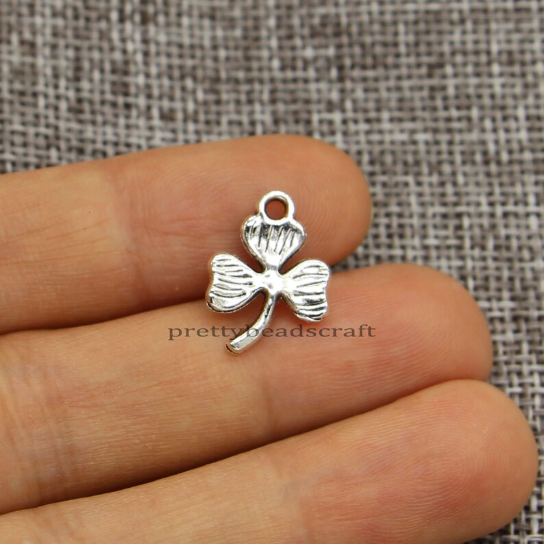 May include: Silver-toned metal charm shaped like a four-leaf clover with a loop at the top for attaching to jewelry or crafts.