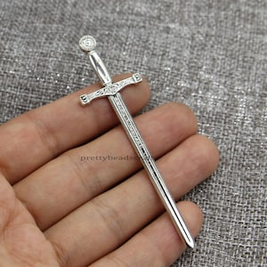 Può includere: Ciondolo a forma di spada argentato con dettagli intricati. La spada ha una punta appuntita e un pomo circolare. Il ciondolo è lungo circa 5 cm.