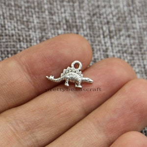 May include: A small, silver Stegosaurus charm with a loop for attaching to jewellery or crafts. The dinosaur has detailed ridges along its back and a textured body. The charm is shown against a neutral background.