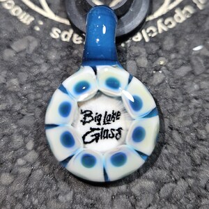 May include: A white glass pendant with a blue top and a blue and white swirl design. The pendant has the words "Big Lake Glass" written in black on the front.
