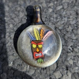 May include: A glass pendant with a gold-colored top and a clear round bottom. The pendant features a cartoon image of a tiki mask with a red face, yellow eyes, and a green and yellow headdress.
