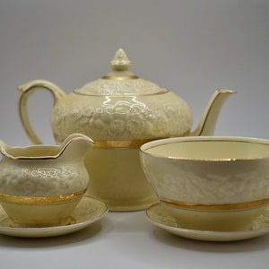 Pode incluir: Um conjunto de chá de cor creme com guarnições douradas. O conjunto inclui uma chaleira, um jarro de creme, um açucareiro e pires. A chaleira tem um design floral.
