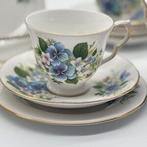 May include: A white teacup and saucer set with a floral pattern featuring blue, pink, and green flowers. The set has a gold trim around the edges.