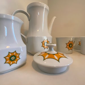 Może przedstawiać: Biały ceramiczny zestaw do herbaty z zielonym i pomarańczowym wzorem kwiatowym. Zestaw zawiera czajnik, dzbanek na śmietanę, cukiernicę z pokrywką i dwie filiżanki.