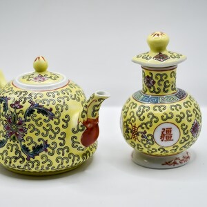 May include: A yellow teapot and a yellow jug with intricate floral and geometric designs. Both pieces feature a red character in a circle on the front.