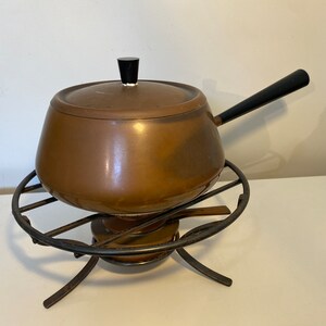 May include: A copper-colored fondue pot with a black handle and lid knob, resting on a black metal stand. The pot has a rounded shape and a lid. The stand has a circular design with legs for stability. A classic piece for entertaining.