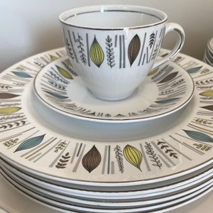 May include: A set of white ceramic plates with a stylised leaf and stem design in shades of brown, green, and blue. The plates have a gold trim around the edges.