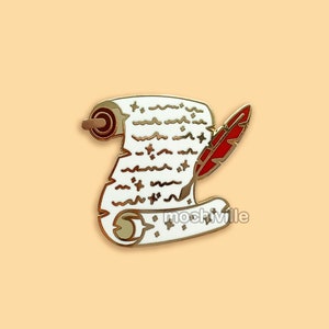 May include: A gold-coloured enamel pin depicting a scroll with white paper and brown edges. A red feather quill is positioned beside the scroll. The pin has a white background and is set against a light orange background.