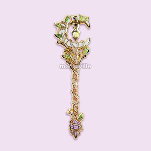 May include: Gold-tone metal pin with a crescent moon design, a lantern, and a crystal at the bottom. The pin is decorated with green leaves and brown branches.