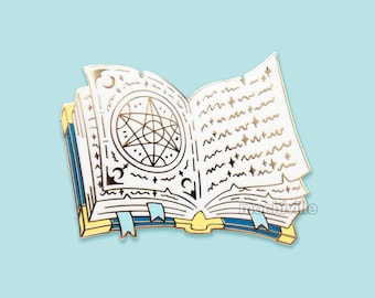 May include: A gold-toned enamel pin of an open book with a five-pointed star inside a circle on the left page. The book is white with black and blue details. The pin is a whimsical and magical design.