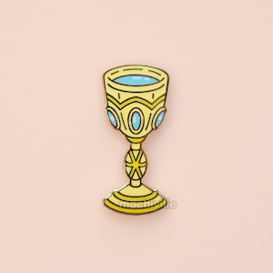 May include: A yellow enamel pin in the shape of a chalice. The chalice has a blue interior and is decorated with gold and blue accents. The pin has a starburst design on the stem and a rounded base. The background is a light pink color.
