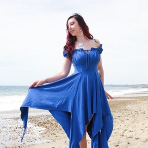 May include: A woman wearing a royal blue off-the-shoulder dress with a flowing skirt and long ties. The dress has a cinched waist and a ruffled neckline. She is standing on a sandy beach with the ocean in the background.