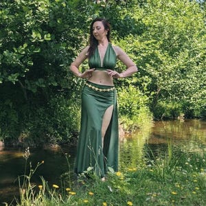 May include: A two-piece olive green outfit consisting of a sleeveless halter-neck crop top with a deep V-neckline and a matching floor-length maxi skirt featuring a high side slit and a decorative gold-toned coin chain belt at the waist.