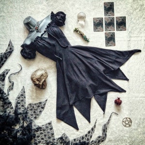 Peut inclure: Une robe gris foncé au design unique et fluide, présentée sur un mannequin. La robe présente un col à volants et une jupe évasée avec des bords pointus. Les accessoires incluent un crâne, des cartes de tarot et un pentagramme.