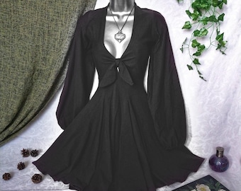 Pode incluir: Um vestido preto com mangas compridas e volumosas e um decote profundo amarrado com um laço. O vestido é exibido em um manequim e apresenta uma saia rodada. Um pingente prateado pende de um cordão preto.