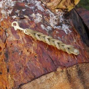 Op de afbeelding: Een gevlochten leren sleutelhanger met een messing haak en een metalen knoop. De sleutelhanger ligt op een stuk hout.