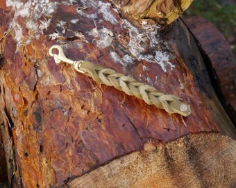 May include: A braided leather key chain with a brass hook and a metal button. The key chain is lying on a piece of wood.