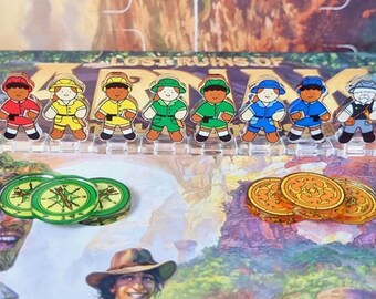 May include: A set of ten colorful explorer figurines, each with a different hat and outfit, stand on a clear plastic base. The figurines are arranged in a row on a game board with the text "LOST RUINS OF ARKA" visible. There are also three green compass tokens and three gold coins on the board.