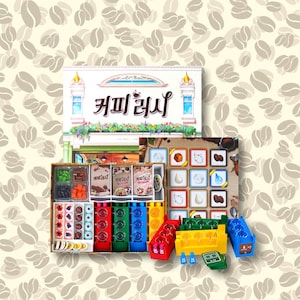 Könnte beinhalten: Ein Brettspiel namens "Coffee Break" mit einem farbenfrohen Spielbrett, bunten Spielsteinen und einer Schachtel mit einer Kaffeeladen-Illustration. Das Spielbrett zeigt verschiedene Bilder, darunter Kaffeebohnen, Tassen und Gebäck.