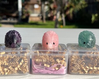 Puede incluir: Cinco pequeñas figuras con forma de erizo en varios colores, incluyendo morado, rosa y verde, se exhiben en cajas de plástico transparente. Las cajas están llenas de papel decorativo y están dispuestas sobre una superficie blanca.