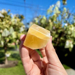 May include: A hand holds a faceted, golden-yellow calcite crystal against a blurred background of green foliage and a blue sky. The crystal has a translucent quality, with visible internal patterns and a white edge.