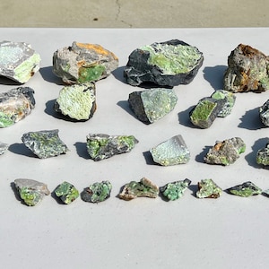 May include: A collection of 25 green and gray rocks with a variety of shapes and sizes. The rocks are arranged on a white surface.