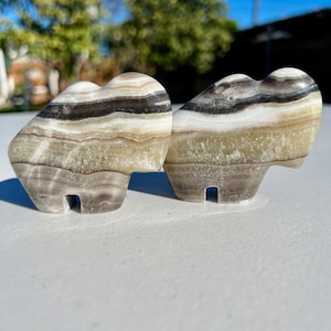 Op de afbeelding: Twee gegraveerde hartvormige sculpturen gemaakt van wit, bruin en grijs gebandeerd onyx. De sculpturen staan ​​tegenover elkaar.