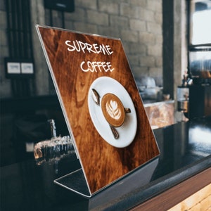 Puede incluir: Un letrero de acrílico transparente con un fondo de madera marrón. El letrero dice "Supreme Coffee" en texto escrito a mano blanco. Una taza de café blanca con arte latte está sobre la superficie de madera.