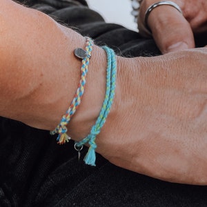 Puede incluir: Una muñeca con tres pulseras, una pulsera de plata, una pulsera de cuentas blancas y una pulsera trenzada con un dije de plata. La persona también lleva un anillo de plata en su dedo.