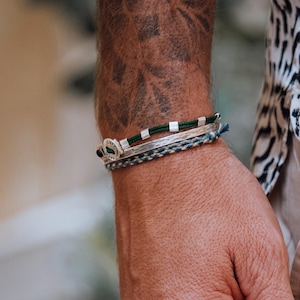 Puede incluir: Una muñeca adornada con tres pulseras. Una es plateada con detalles verdes y blancos, otra es un cordón trenzado azul y plateado, y la tercera es una banda plateada con un diseño verde y negro. La persona lleva una camisa con estampado en blanco y negro.