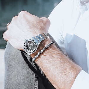 May include: A silver wristwatch with a black face and a black beaded bracelet on a person's wrist.