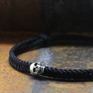 Op de afbeelding: Een zwarte gevlochten touw armband met een zilveren schedel bedel.