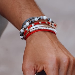 May include: A wrist displaying a collection of bracelets. The bracelets feature diverse designs, including beaded, braided, and corded styles, in red, white, grey, and black. The person is also holding sunglasses.