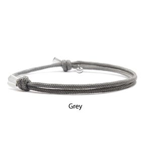 May include: A grey cord bracelet featuring a double-strand design with two adjustable sliding knots. The bracelet includes silver-toned metal end caps and a small circular metal charm attached near one knot. The word Grey is printed below.