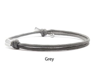 May include: A grey cord bracelet featuring a double-strand design with two adjustable sliding knots. The bracelet includes silver-toned metal end caps and a small circular metal charm attached near one knot. The word Grey is printed below.