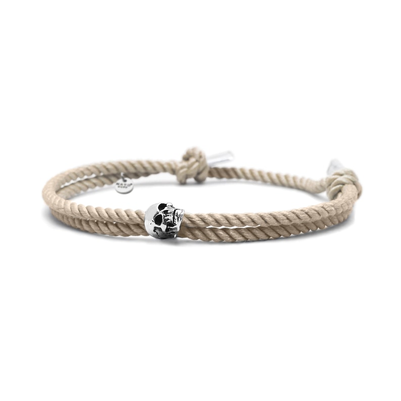 May include: A beige rope bracelet with a silver skull charm. The bracelet is tied with a knot and has a small silver tag.