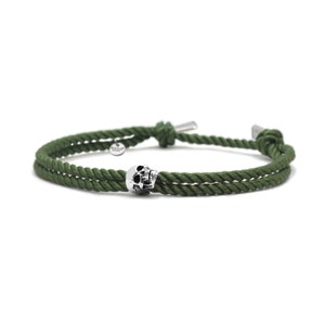 May include: A green rope bracelet with a silver skull charm. The bracelet is made of two strands of rope that are tied together with a knot.