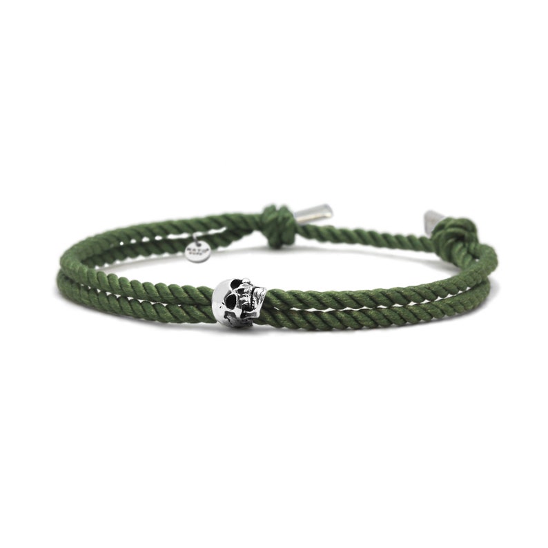 May include: A green rope bracelet with a silver skull charm. The bracelet is made of two strands of rope that are tied together with a knot.