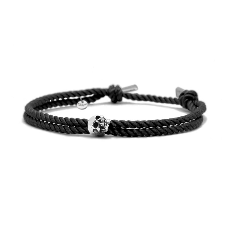 May include: A black rope bracelet with a silver skull charm. The bracelet is tied with a knot on each end.
