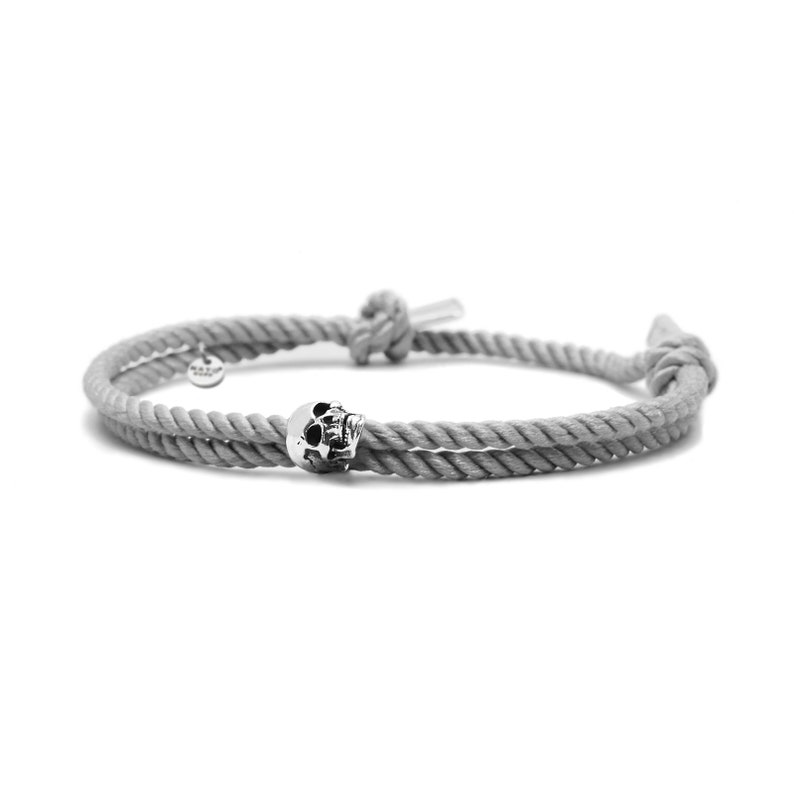 May include: A gray rope bracelet with a silver skull charm. The bracelet is tied with a knot on one end and has a small silver tag on the other end.