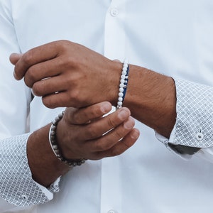 Op de afbeelding: Een witte kralen armband met een blauw leren bandje om de pols van een persoon. De armband wordt gedragen met een wit overhemd met knopen en een gepatenteerde manchet.