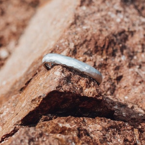 Puede incluir: Una alianza de boda de plata martillada descansando sobre una superficie rocosa marrón y rugosa.