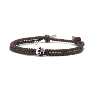May include: A brown rope bracelet with a silver skull charm. The bracelet is tied with a knot on each end.