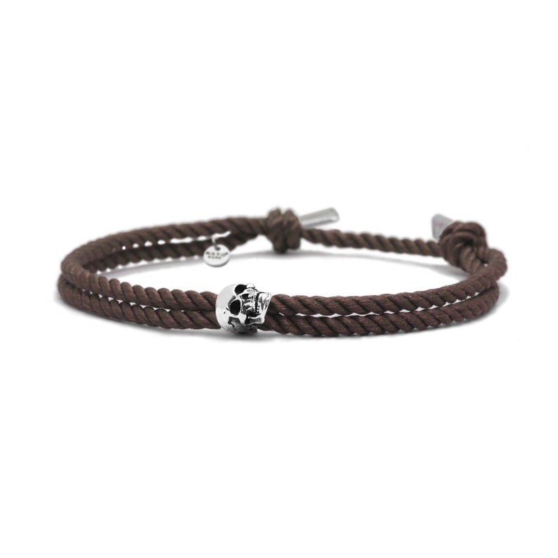 May include: A brown rope bracelet with a silver skull charm. The bracelet is tied with a knot on each end.