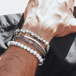 May include: A man's wrist wearing four bracelets. The bottom bracelet is white with round beads. The next bracelet is a gray rope bracelet with a silver skull charm. The third bracelet is a silver bangle. The top bracelet is made of silver hexagonal beads.