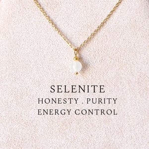 Puede incluir: Un collar de cadena dorada con un colgante de una pequeña cuenta redonda de selenita blanca. Debajo del colgante, el texto dice: SELENITE, HONESTY . PURITY, ENERGY CONTROL. El collar se muestra sobre un soporte de tela beige claro ovalado.