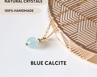 May include: A gold-toned chain necklace featuring a single round, light blue calcite bead pendant. The image includes the text NATURAL CRYSTALS, 100% HANDMADE, and BLUE CALCITE. The jewelry is displayed near a gold-colored geometric object.