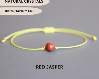 May include: A bracelet featuring a single round red jasper bead centered on a thin yellow cord. The bead is flanked by two small gold-toned metal spacers. Text on the image reads NATURAL CRYSTALS, 100% HANDMADE, and RED JASPER.