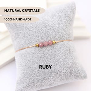 Puede incluir: Un brazalete delicado con una piedra preciosa de rubí natural y detalles dorados. El brazalete se muestra en un cojín de terciopelo gris con el texto "NATURAL CRYSTALS 100% HANDMADE" y "RUBY" impreso en él.