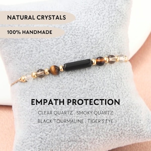 Può includere: Un braccialetto di perline con un cilindro centrale in tormalina nera, affiancato da perline di occhio di tigre, quarzo fumé e quarzo chiaro con distanziatori dorati. Testo: NATURAL CRYSTALS, 100% HANDMADE, EMPATH PROTECTION, CLEAR QUARTZ, SMOKY QUARTZ, BLACK TOURMALINE, TIGER'S EYE.
